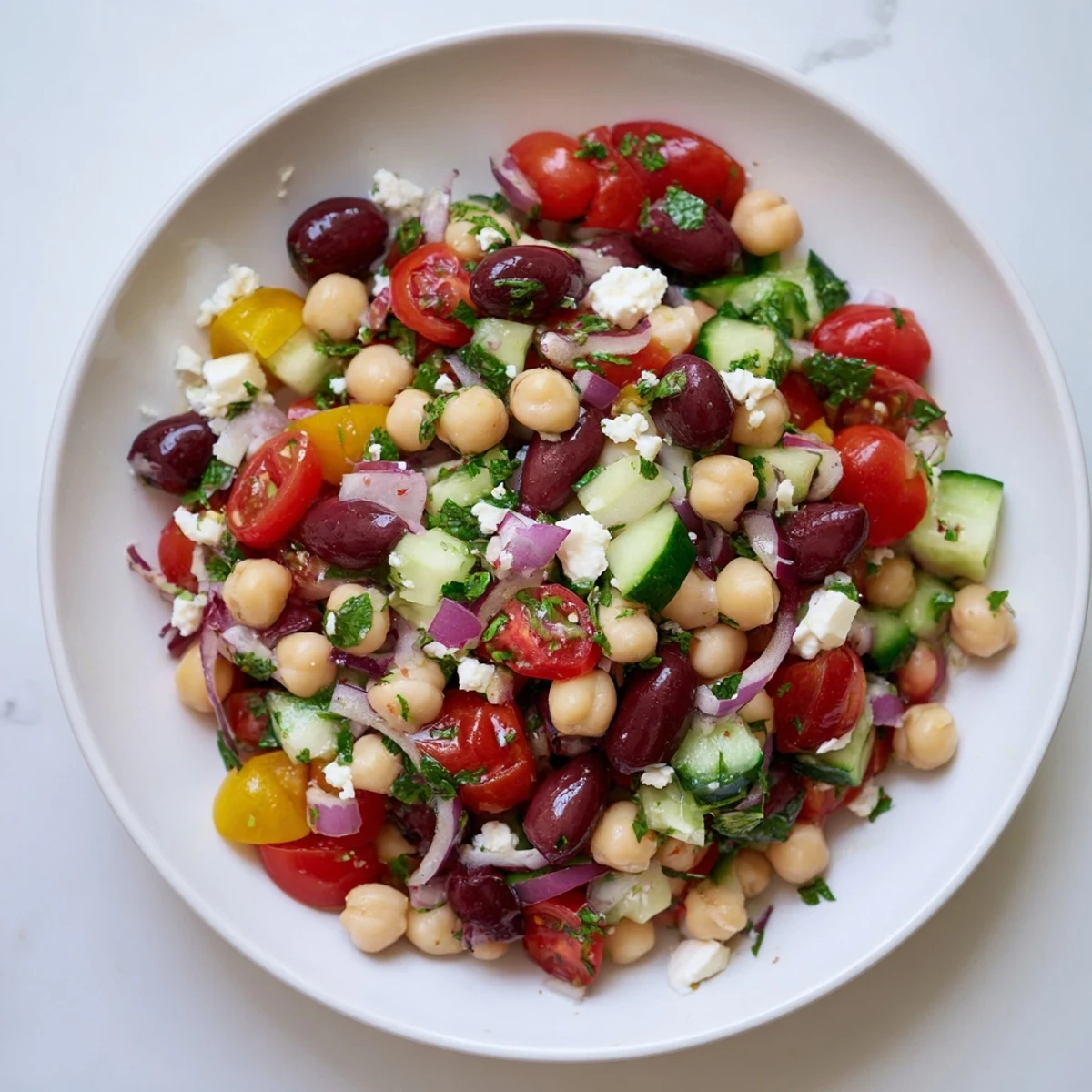 A close-up of Mediterranean Dense Bean Salad with chickpeas, tomatoes, and feta in a white bowl, garnished with fresh parsley.