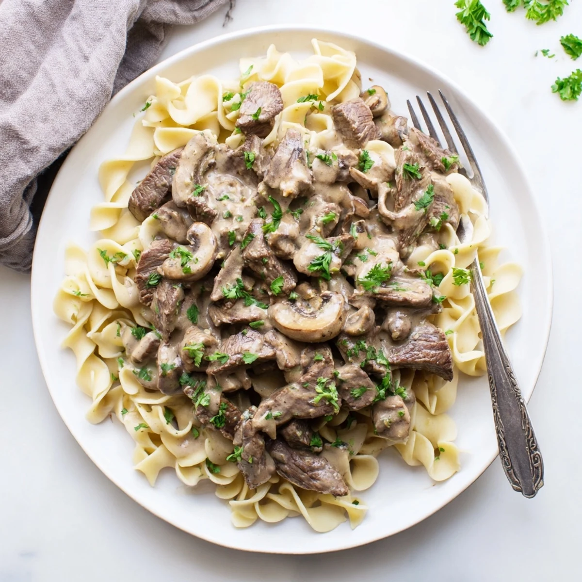 Homemade beef and mushroom stroganoff plated over buttery noodles with chopped parsley sprinkled on top