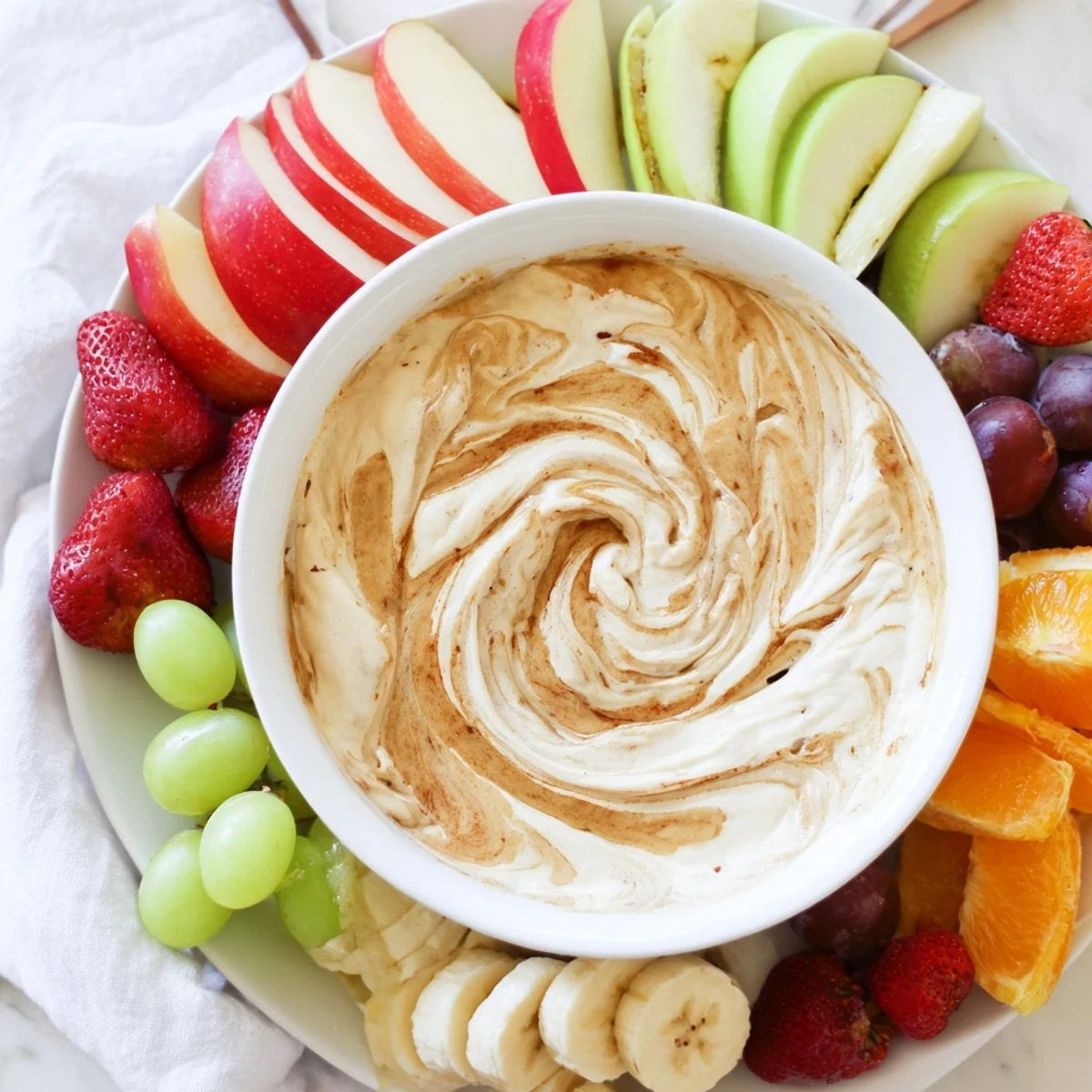 Healthy cinnamon roll dip made with Greek yogurt and cream cheese, topped with sweet cinnamon swirl and served with crisp fruit dippers on a white platter